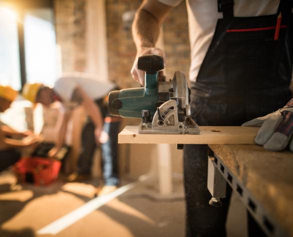 Unrecognizable manual worker using circular saw while cutting piece of wood at construction site. His colleagues are in the background.